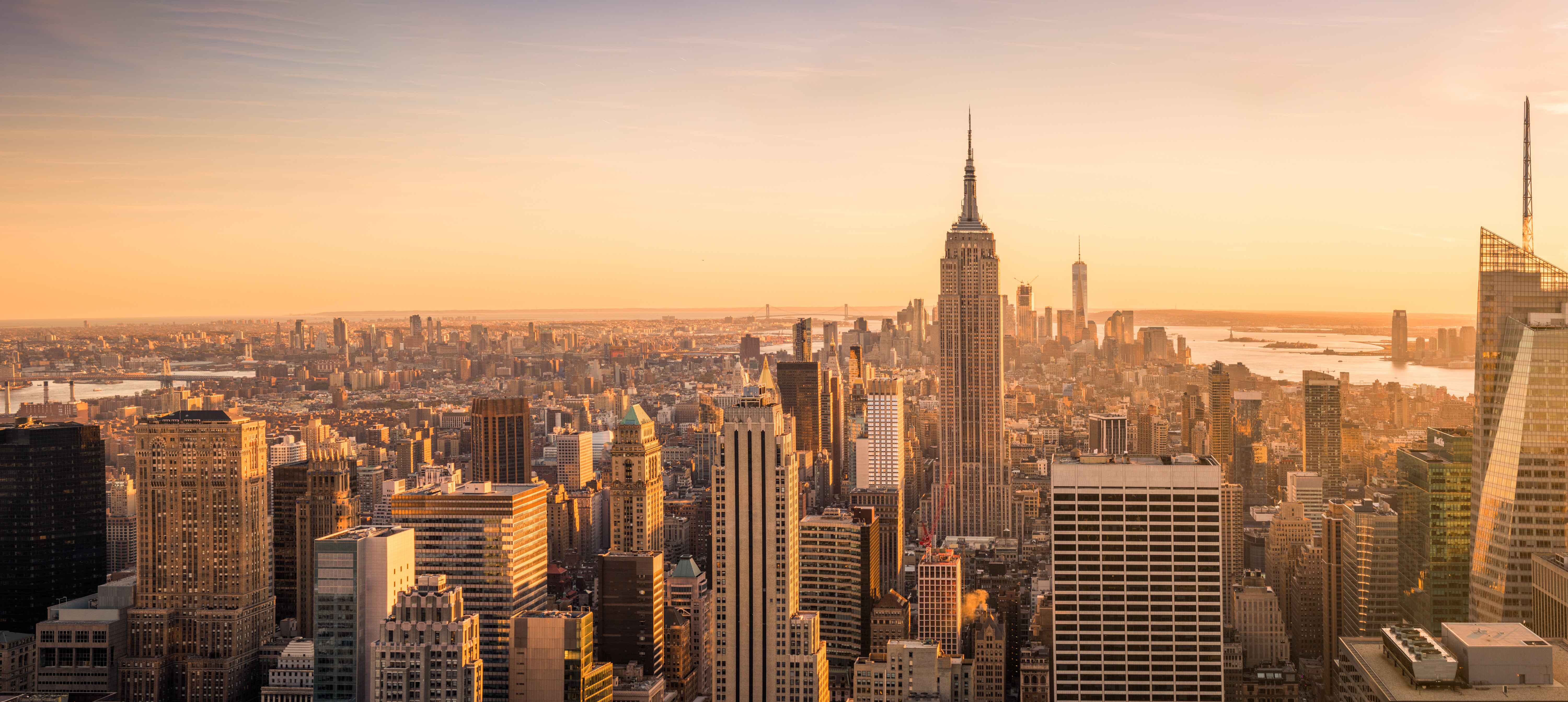 Panoramic view of New York City skyline at sunset with skyscrapers and the Hudson River in the background.