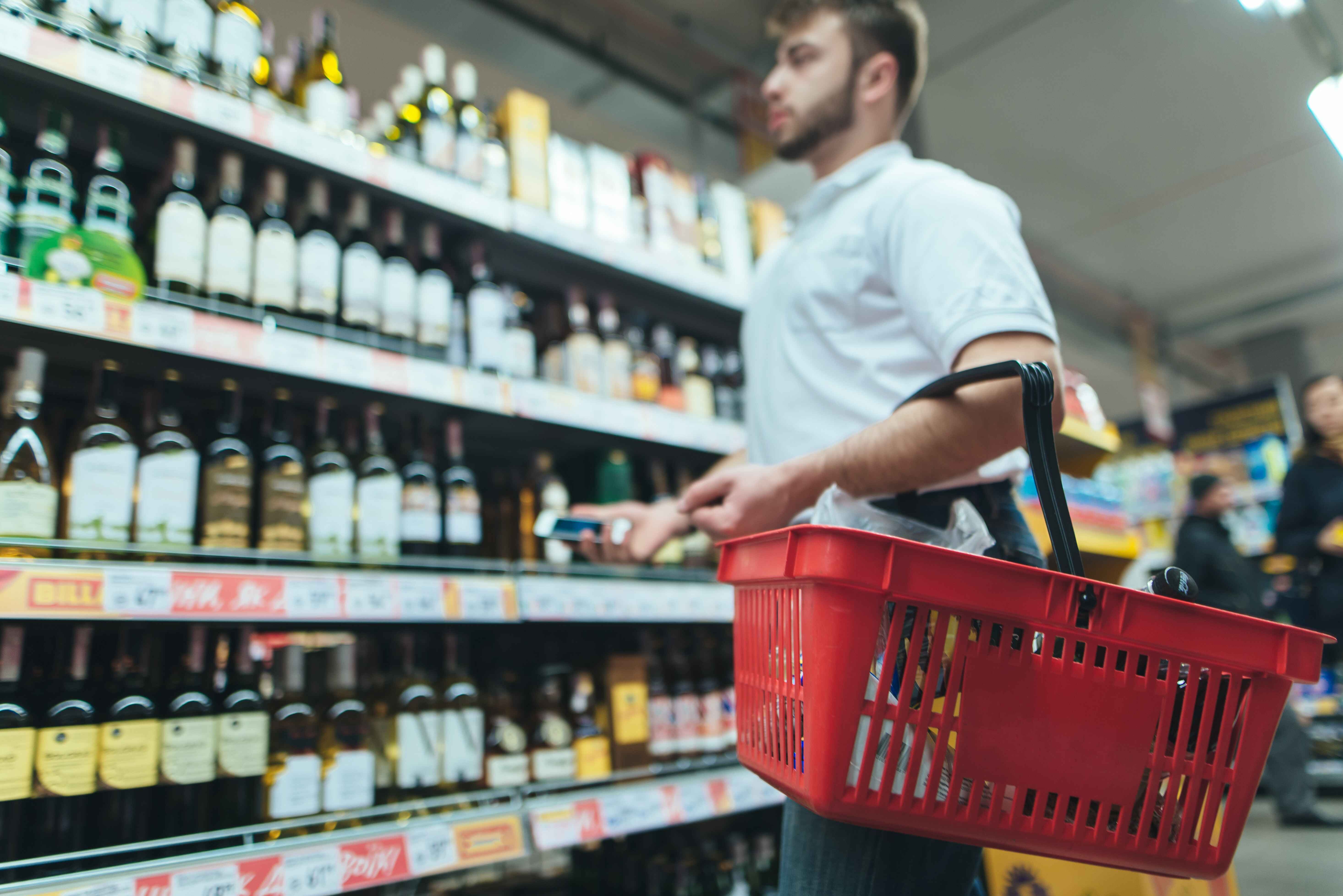 Man shopping for liquor in a grocery store aisle holding a red basket and a smartphone.