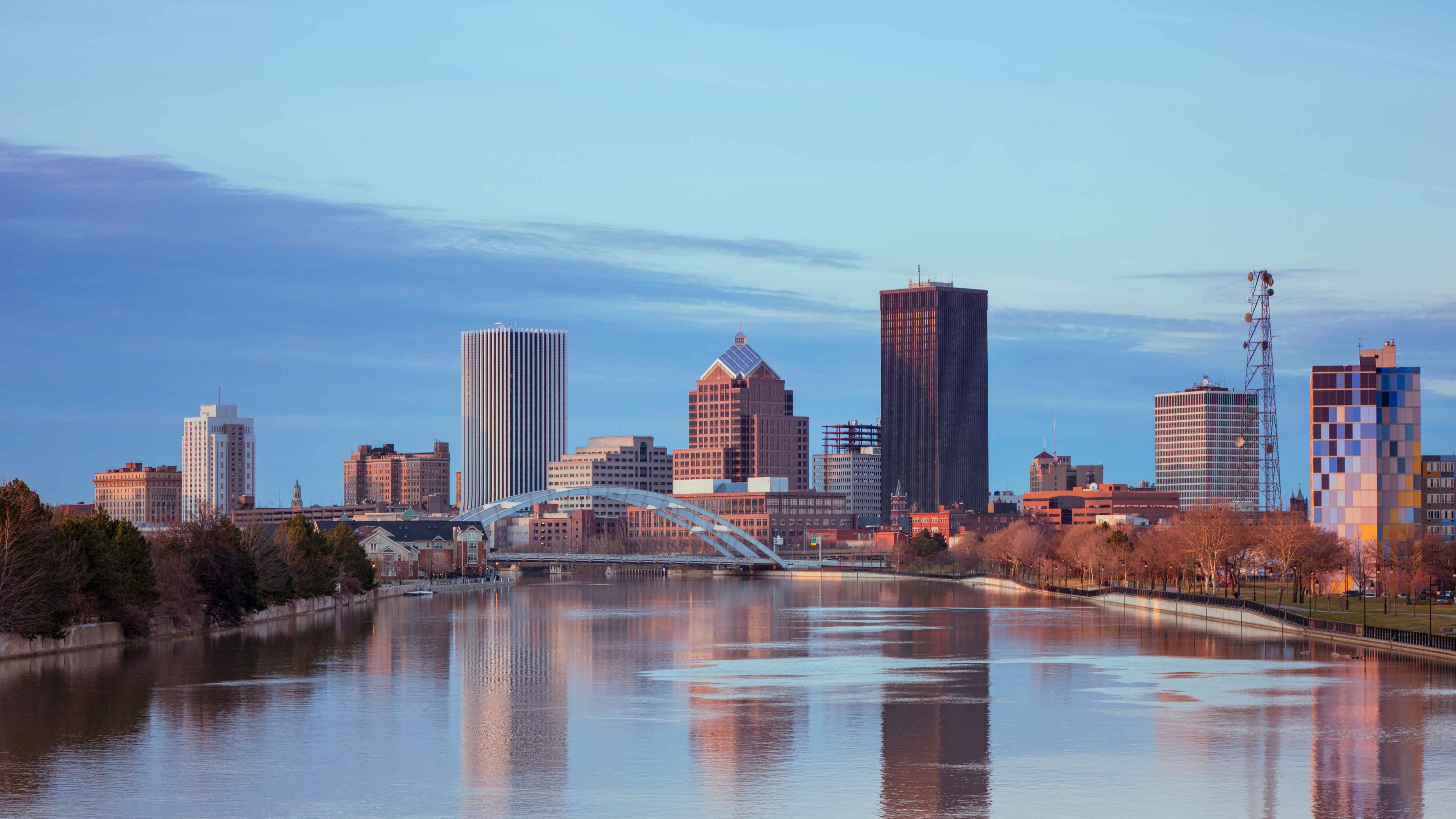 City skyline with skyscrapers reflected in a calm river, featuring an arched bridge and a sunset sky.