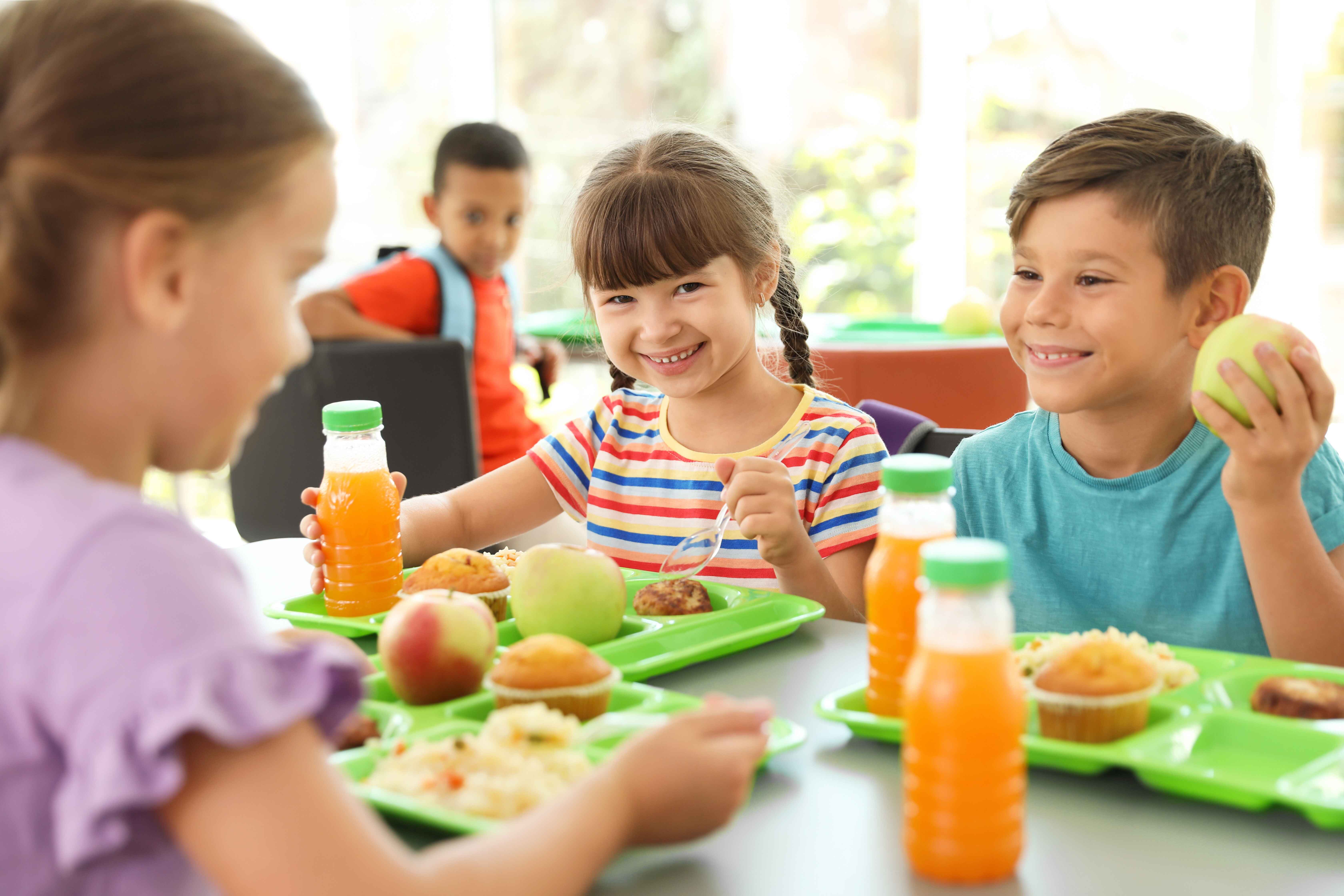 Children smiling and having lunch at a table with green trays, containing orange juice, muffins, and fruit.