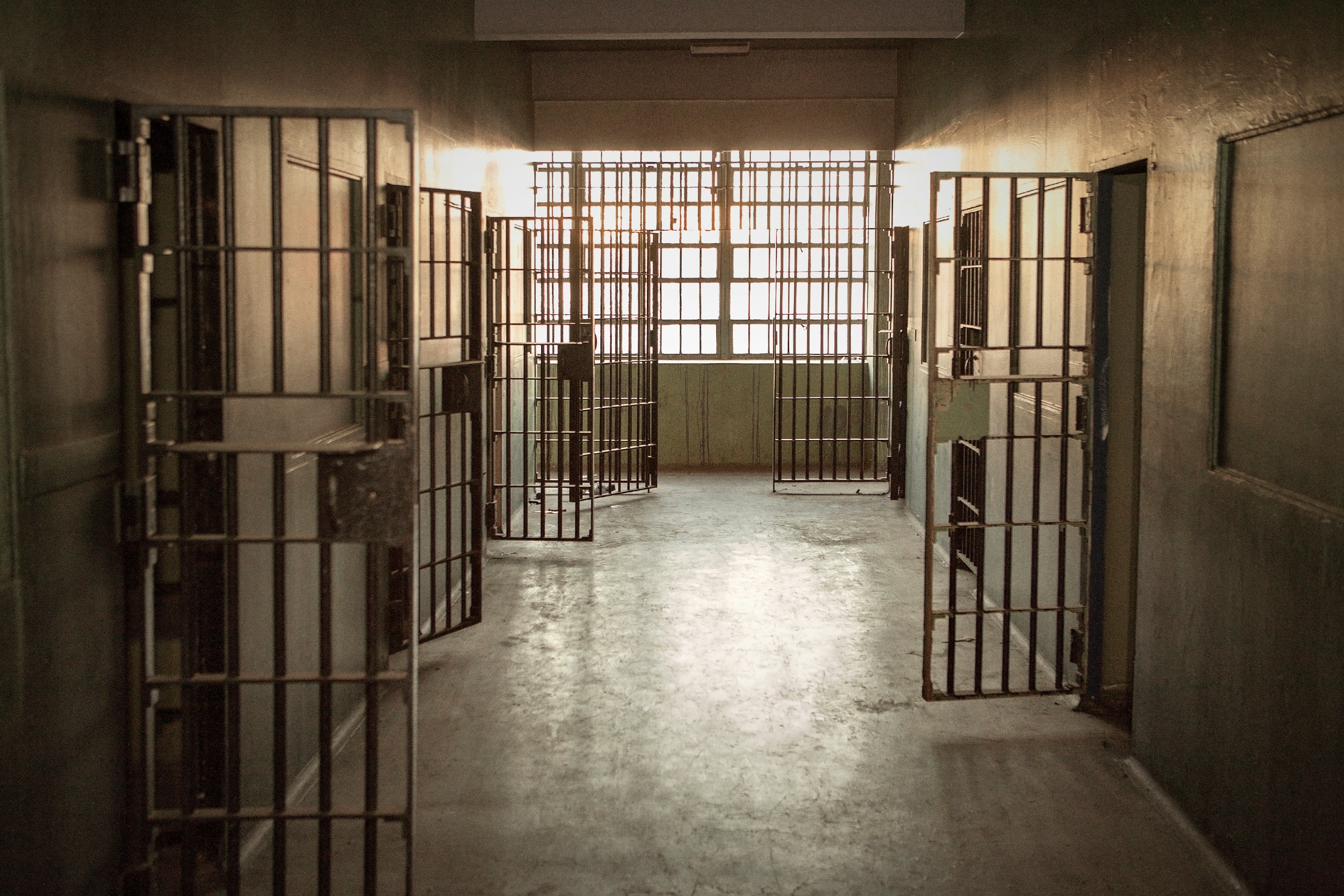 A prison corridor with open barred cell doors and a window at the end illuminating the space.