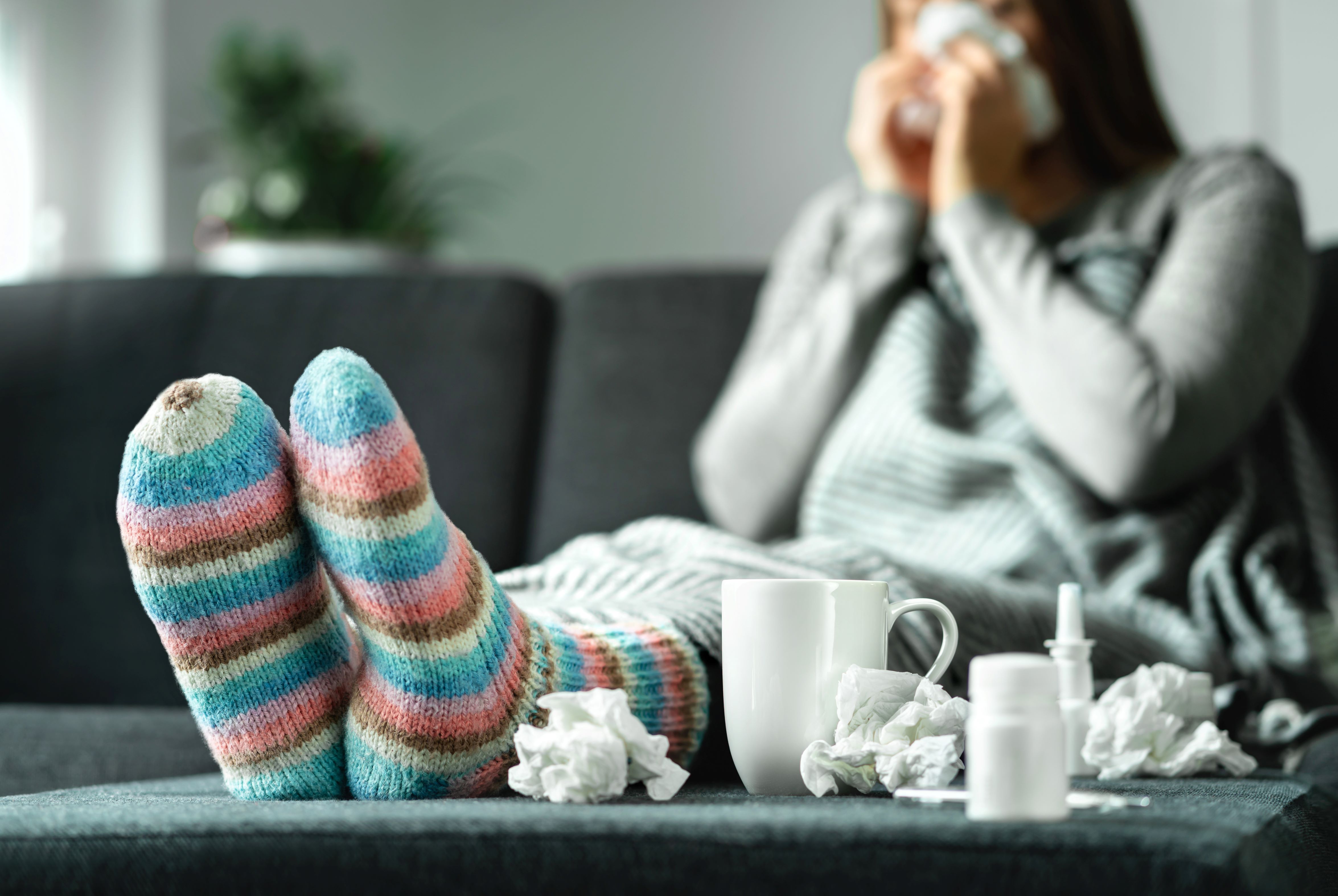 Person sits on a couch with feet up surrounded by tissues and a tea cup