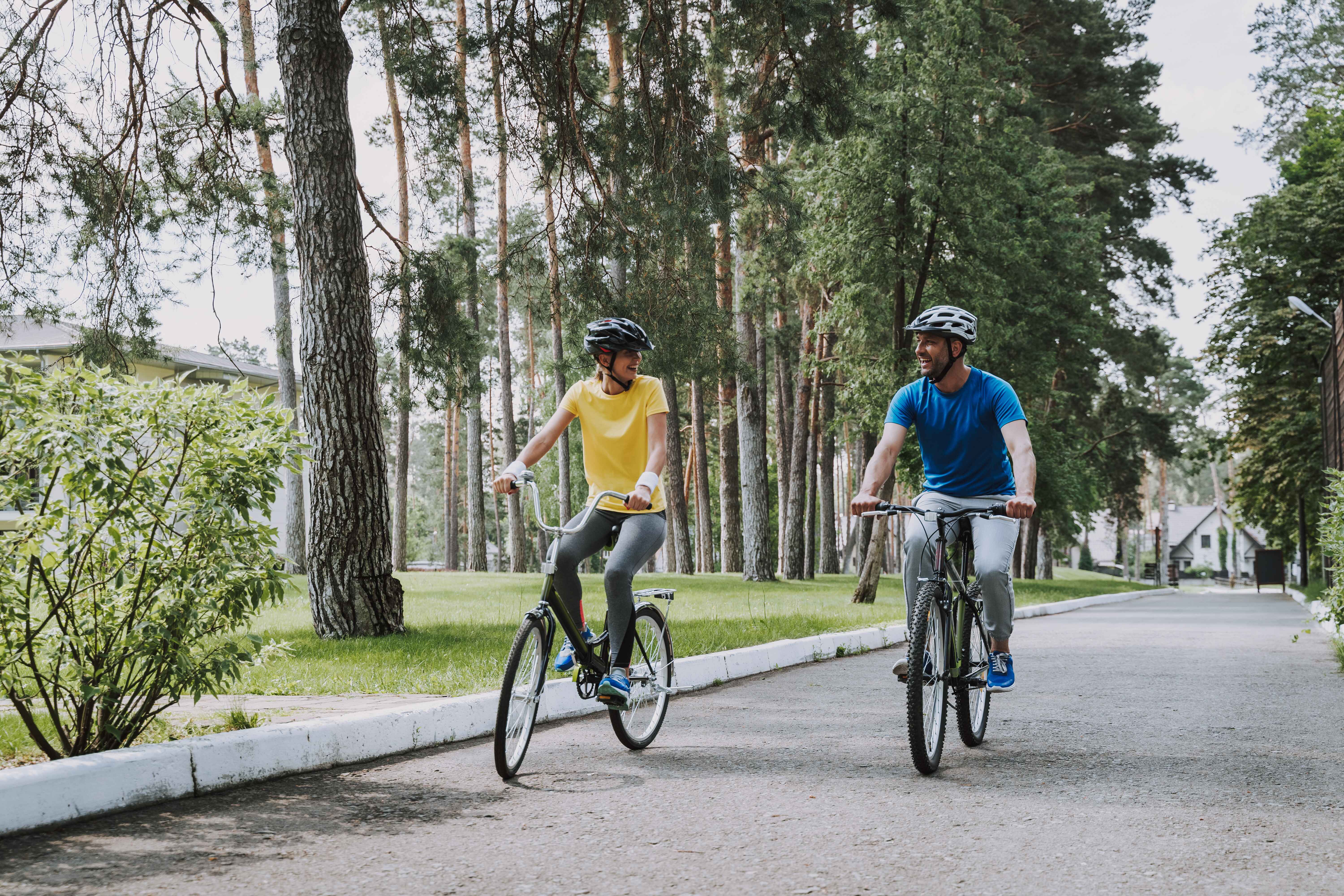 A man and a woman cycling on a park path, surrounded by grass and trees, on a sunny day.