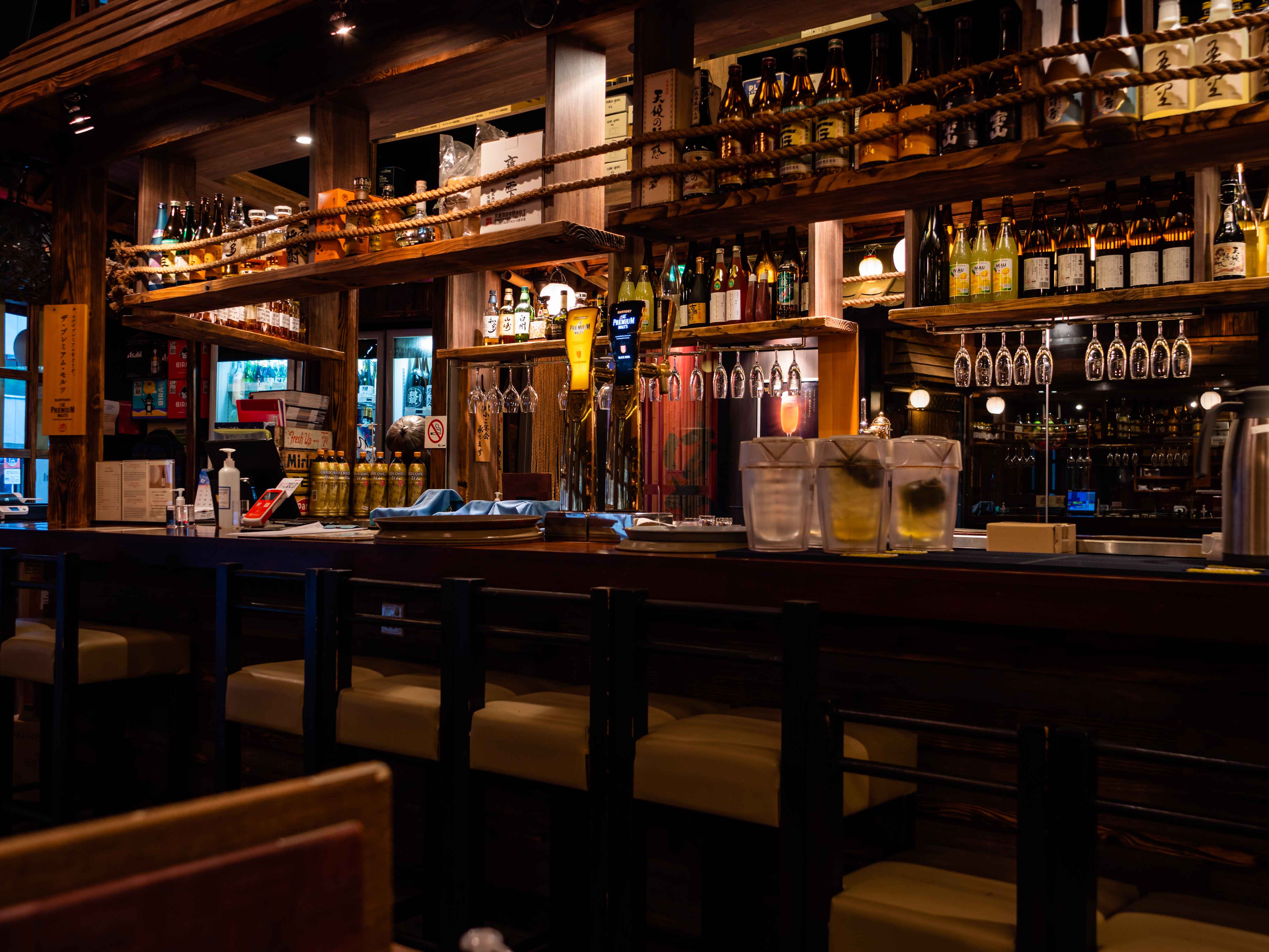 Interior of a bar with shelves of liquor bottles and hanging wine glasses, with a wooden counter and stools.