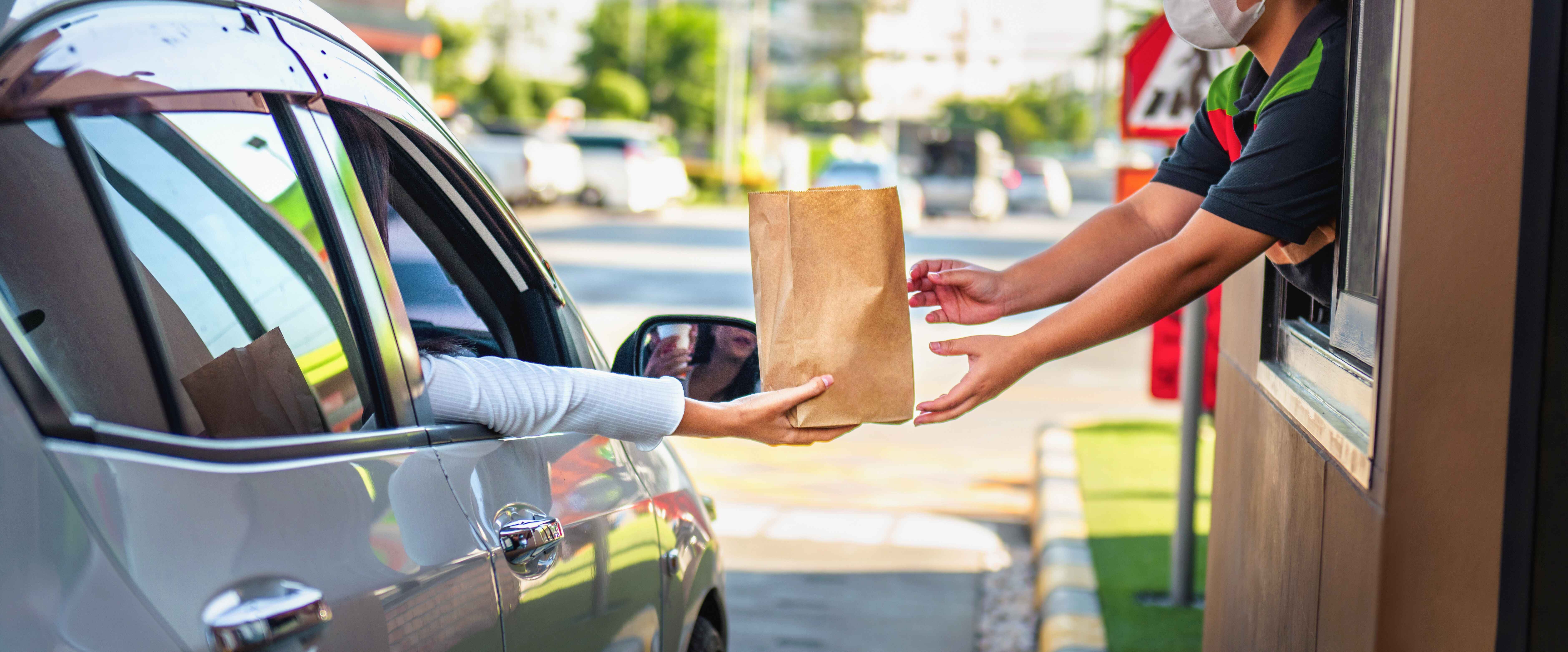 Drive-thru transaction with a person in a silver car receiving a brown paper bag from a masked employee at a window.