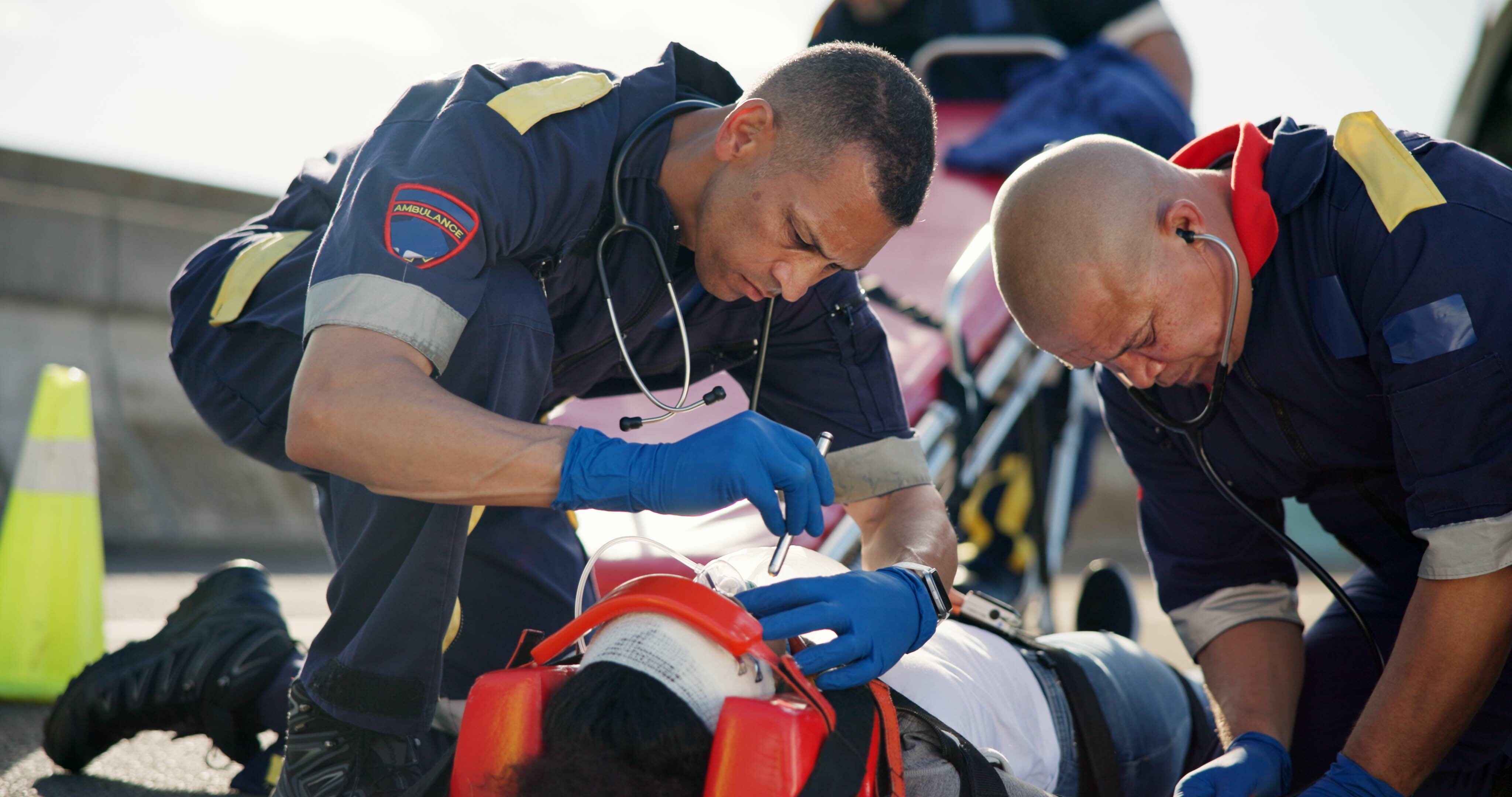 Two paramedics treat a patient with a bandaged head and oxygen mask on the ground.