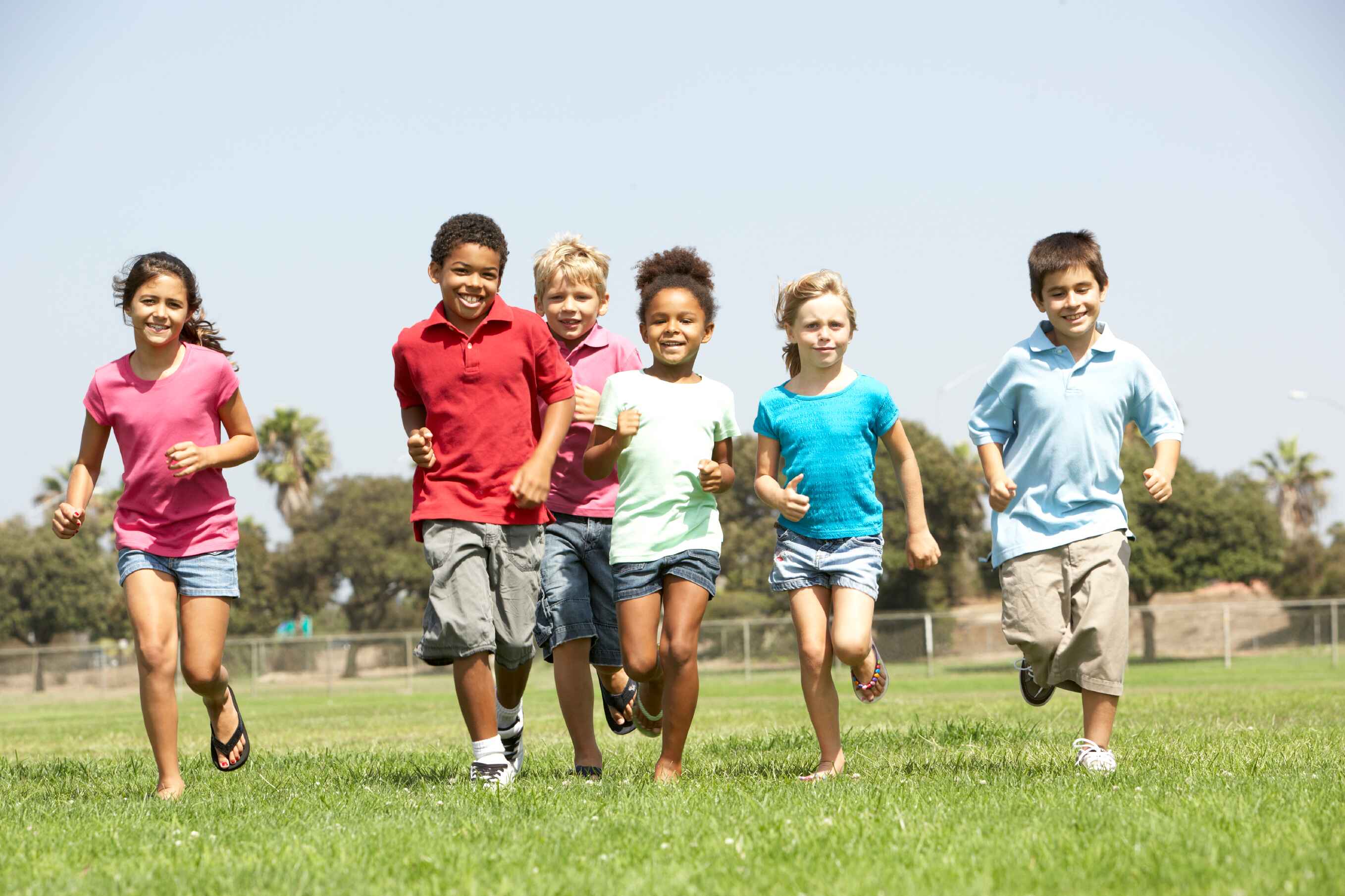 Six children running on grass in a park under a clear sky.