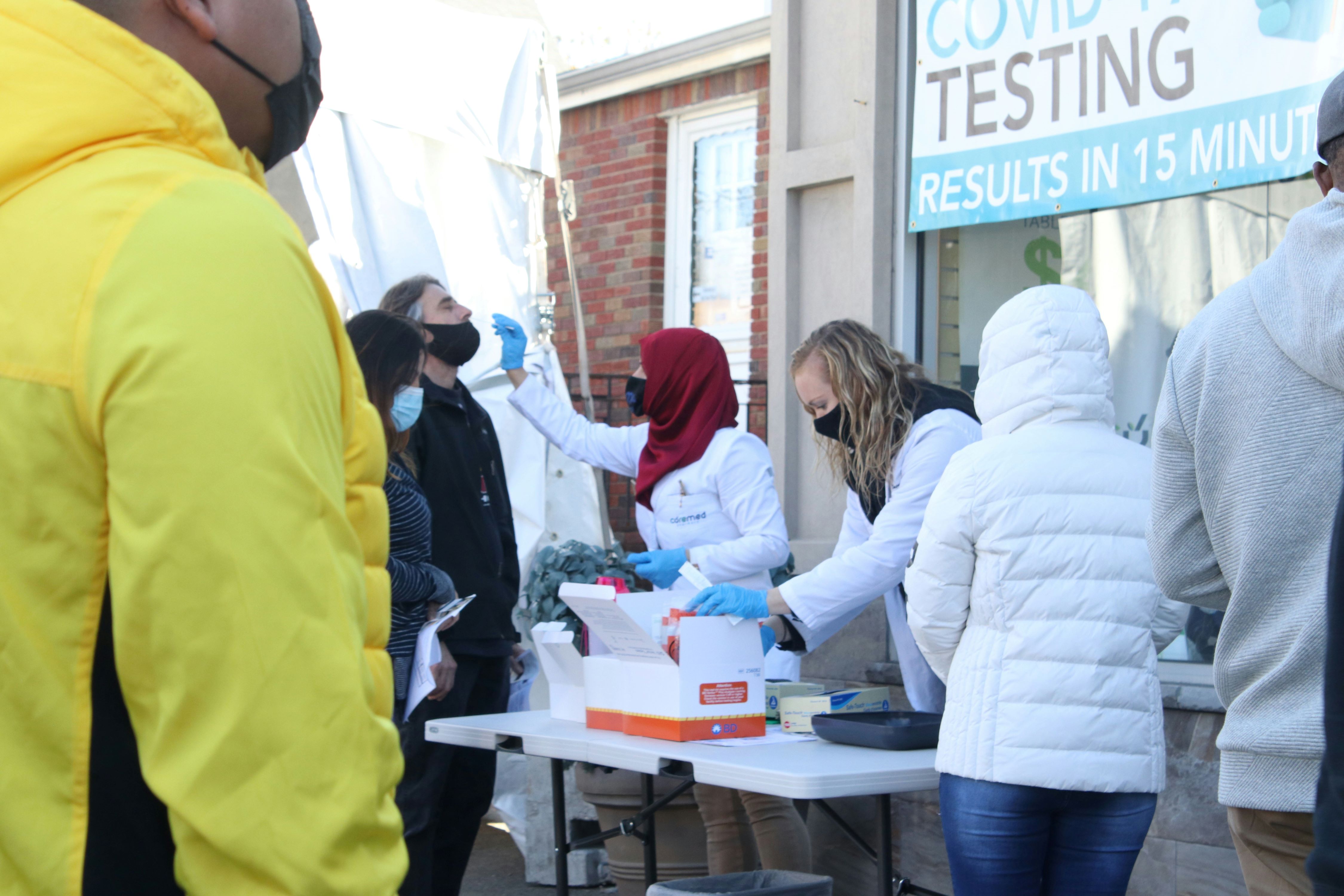 Outdoor COVID-19 testing site with people in line and healthcare workers administering tests.