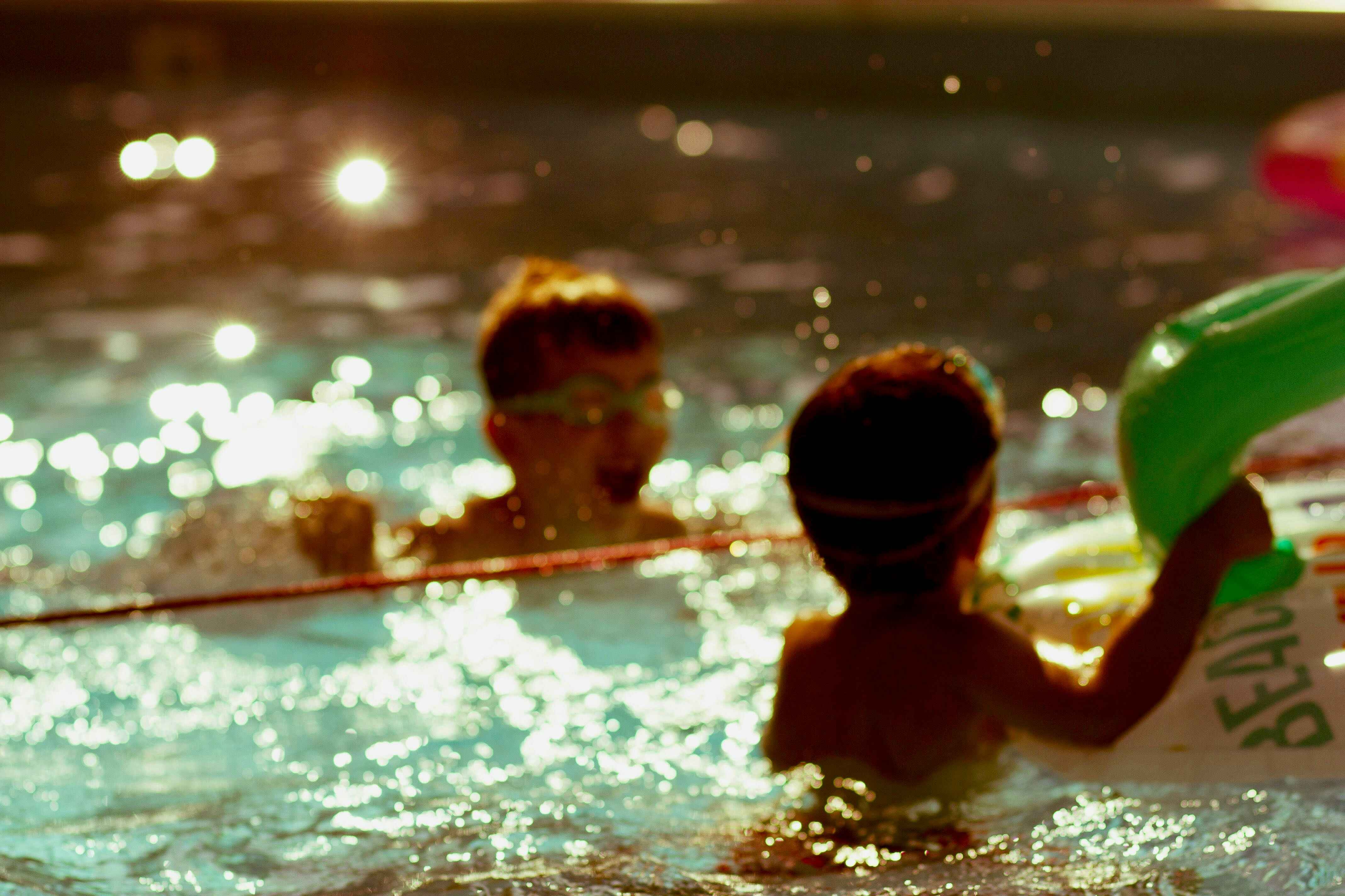 Two children in a sunlit swimming pool, one wearing goggles and another holding a green inflatable float.