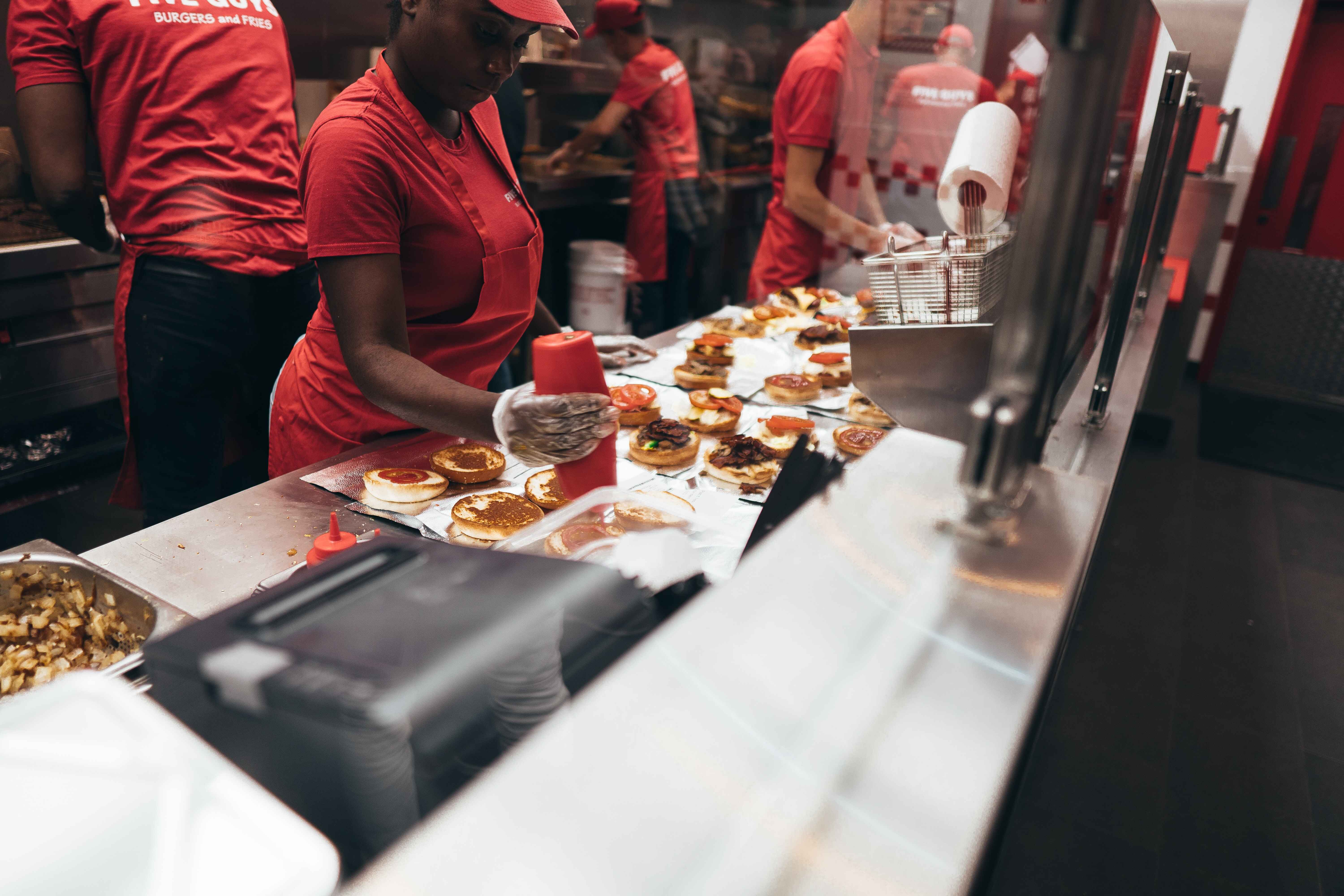 Restaurant kitchen workers assembling burgers, with one applying condiments onto buns.