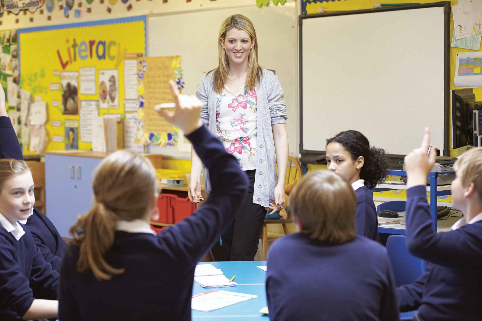 A classroom scene with a teacher and students in blue uniforms, with educational posters and a whiteboard in the background