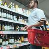 Man shopping for liquor in a grocery store aisle holding a red basket and a smartphone.