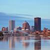 City skyline with skyscrapers reflected in a calm river, featuring an arched bridge and a sunset sky.