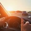 View from inside a car driving on a highway at sunset, with hands on the steering wheel and vehicles ahead.