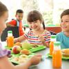 Children smiling and having lunch at a table with green trays, containing orange juice, muffins, and fruit.