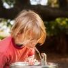 Child drinking from an outdoor water fountain with blurred greenery in the background.