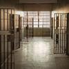 A prison corridor with open barred cell doors and a window at the end illuminating the space.