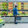 A man with a shopping basket stands in front of a variety of fruits and vegetables in a supermarket.