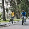 A man and a woman cycling on a park path, surrounded by grass and trees, on a sunny day.
