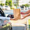 Drive-thru transaction with a person in a silver car receiving a brown paper bag from a masked employee at a window.
