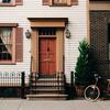 Facade of a traditional house with cream siding, brown shutters, a central entrance, and a red bicycle.