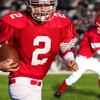 Football player in red jersey holding a football, with another player and a crowd in the background.