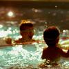 Two children in a sunlit swimming pool, one wearing goggles and another holding a green inflatable float.