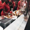 Restaurant kitchen workers assembling burgers, with one applying condiments onto buns.